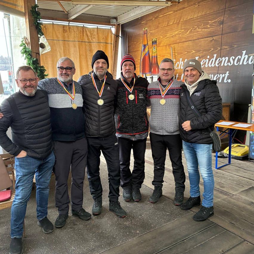 Fünf Männer stehen eng beieinander, tragen Medaillen und lächeln, möglicherweise bei einer Feier. Hinter ihnen ist eine Holzwand mit Text und eine Flasche auf einem Regal.
