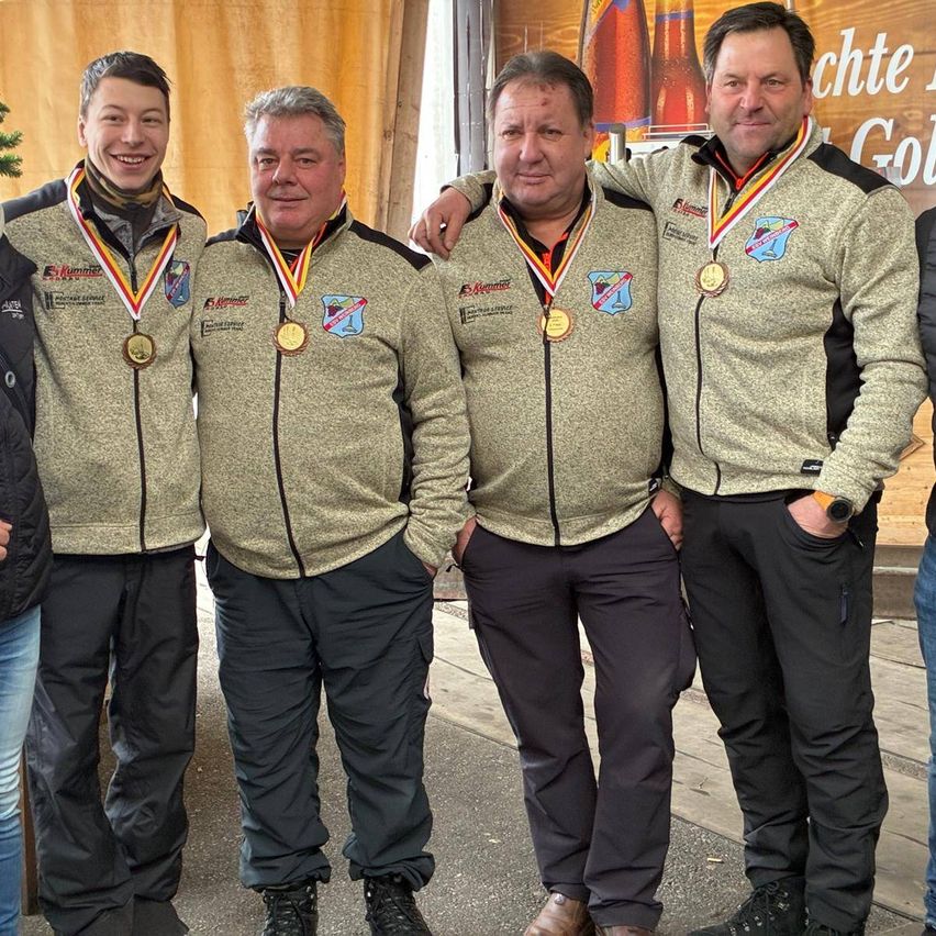 Vier Männer mit Medaillen stehen zusammen, wahrscheinlich bei einer Veranstaltung. Sie tragen verschiedene Jacken, alle mit Logos, und haben Turnschuhe an. Der Mann auf der linken Seite lächelt, während die anderen neutrale Gesichtsausdrücke haben. Hinter ihnen befindet sich ein Banner.