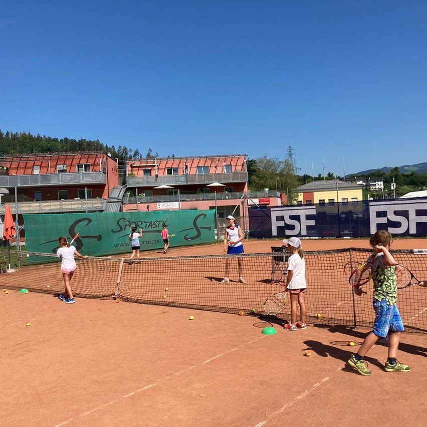 Eine Gruppe von Kindern spielt Tennis auf einem Platz, wobei eine Frau mit einer weißen Mütze beobachtet. Der Platz ist von einem Netz umgeben und hat Tennisbälle, die auf dem Boden verstreut sind. Hinter dem Platz befinden sich Gebäude und Bäume.