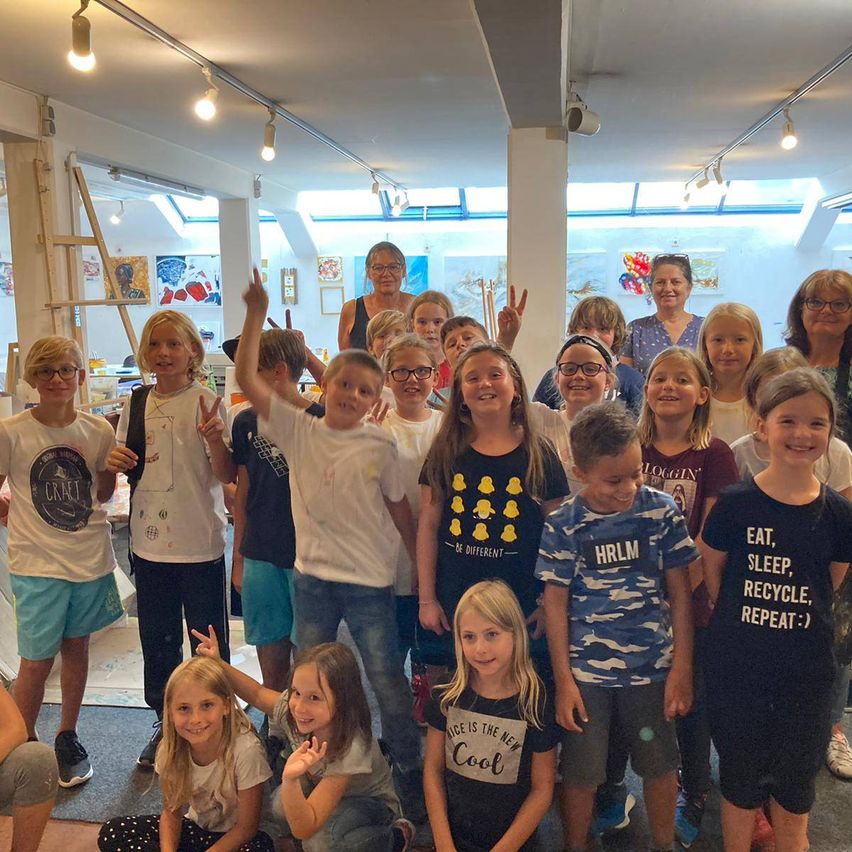 Eine Gruppe von Kindern und Erwachsenen posiert für ein Foto in einem Raum mit weißen Wänden und Glasfenstern. Die Kinder tragen verschiedene Shirts und einige tragen Brillen.