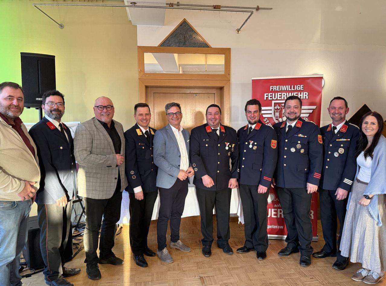 Eine Gruppe von Männern in Uniformen und Anzügen steht in einem Raum und lächelt für ein Foto. Hinter ihnen steht ein Banner mit der Aufschrift 'Freiwillige Feuerwehr'.