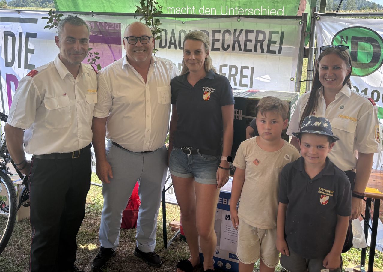 A group of adults and children pose for a photo in front of a banner with the text 'macht den Unterschied'. The woman in the middle wears a black shirt with the logo of the Bundeswehr. The man on the left wears a white shirt and black pants. The man on the right wears a white shirt and gray pants.