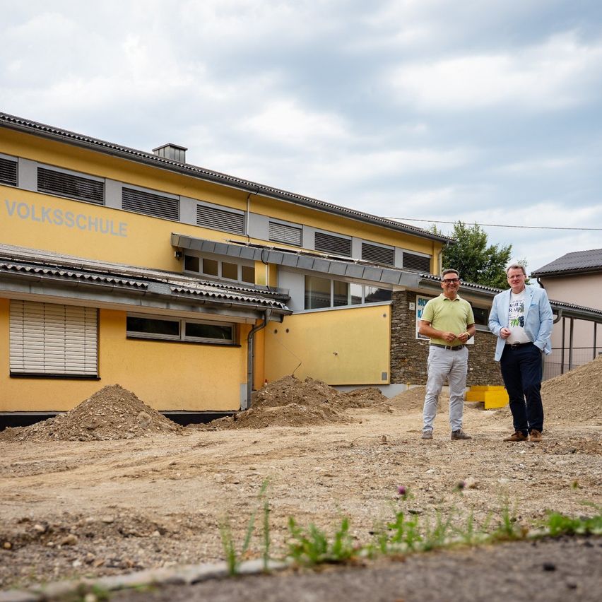 Zwei Männer stehen vor einem gelben Gebäude mit der Aufschrift 'Volksschule'. Sie sind von Erde und Baumaterialien umgeben.