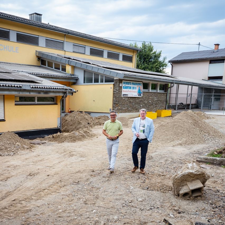 Zwei Männer stehen vor einer Baustelle, einer trägt ein grünes Shirt und der andere eine hellblaue Jacke. Hinter ihnen befindet sich ein Gebäude mit Solarpanelen und einem Banner.