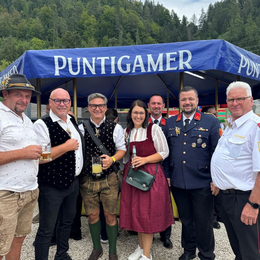 Eine Gruppe von Menschen in traditioneller deutscher Kleidung und Uniformen steht unter einem blauen Zelt mit dem Wort 'Puntigamer'. Sie lächeln und posieren für ein Foto.