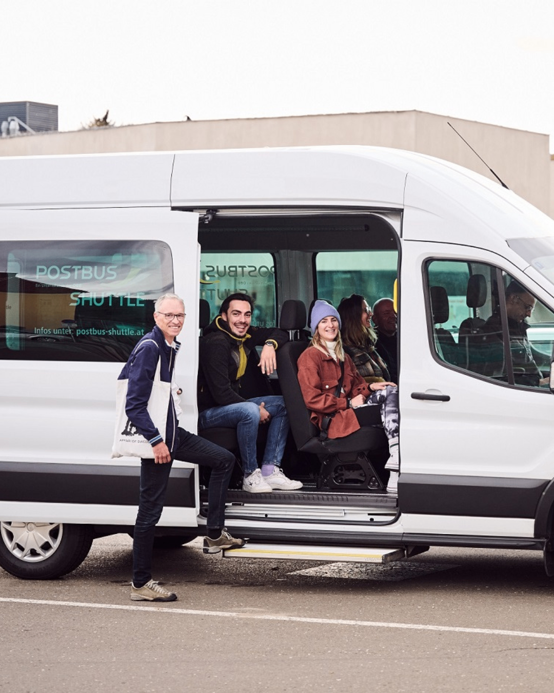 Eine Gruppe von Menschen sitzt in einem weißen Van mit der Aufschrift 'Postbus Shuttle', während ein Mann außerhalb des Vans steht.
