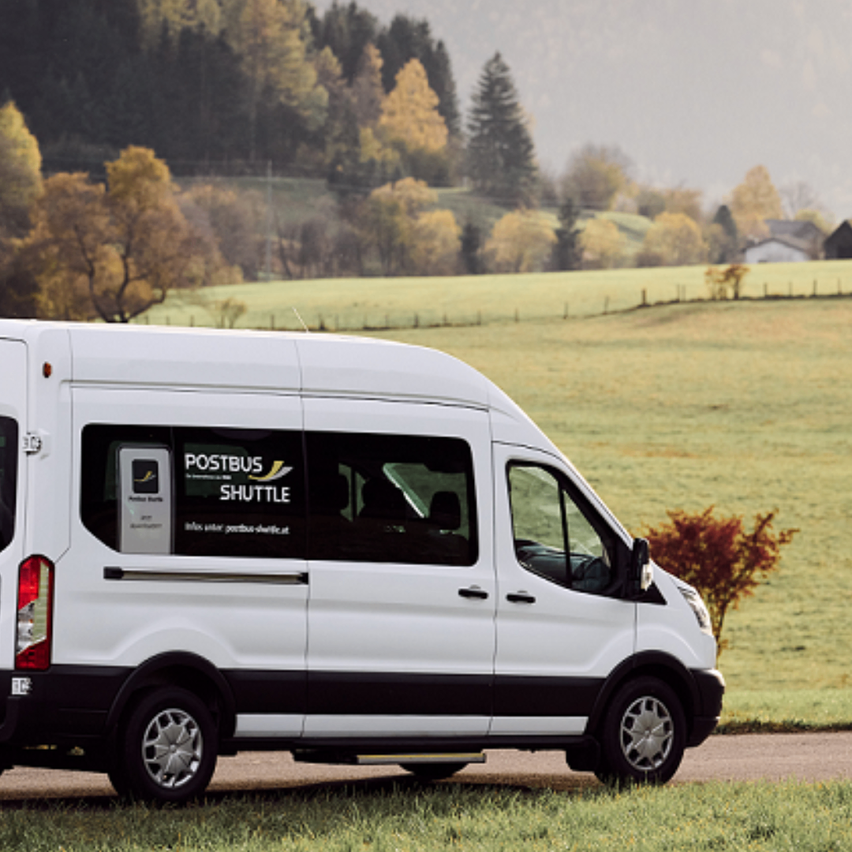 Ein weißer Postbus Shuttle Van steht am Straßenrand auf einer Wiese mit Blick auf ein grünes Feld und Bäume im Hintergrund.