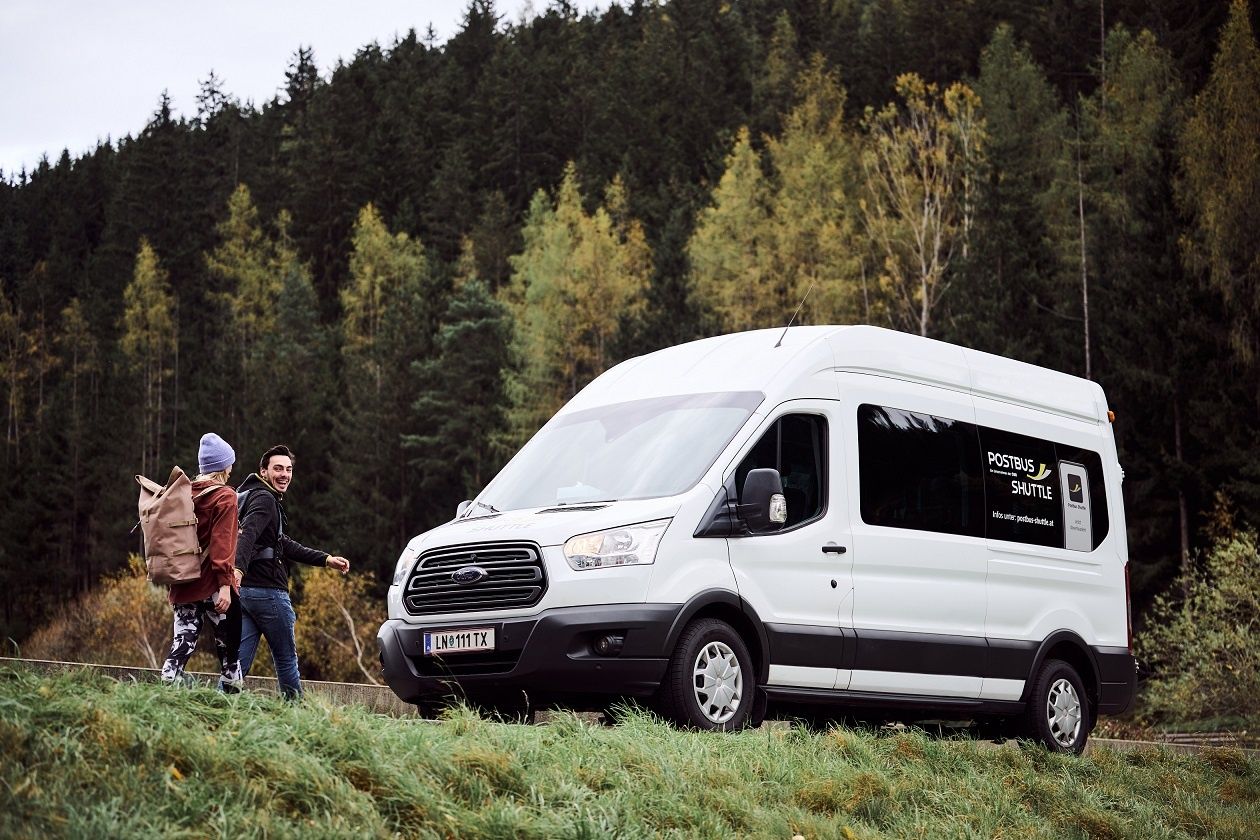 Ein weißer Ford Transit-Transporter mit einem 'Postbus Shuttle'-Schild ist auf einer Wiese geparkt, mit einem Wald im Hintergrund. Zwei Personen gehen auf den Van zu.