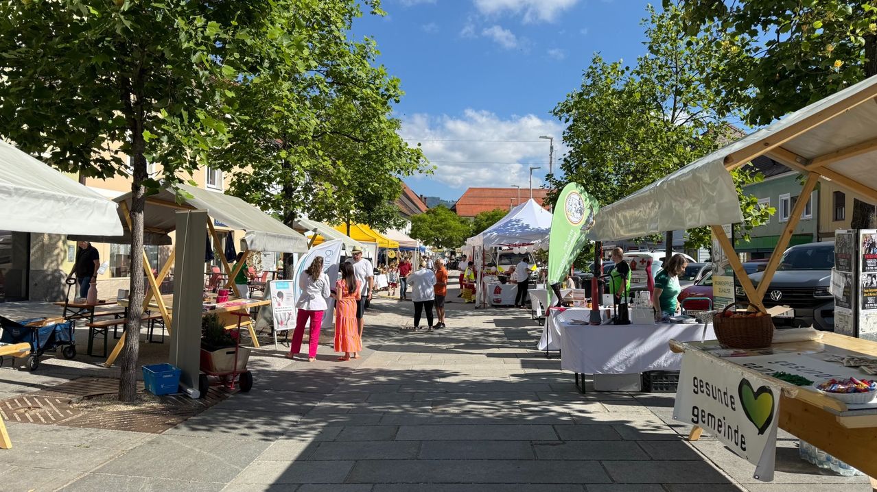 Ein Straßenmarkt mit bunten Zelten und Menschen, die die Stände durchstöbern. Bäume und Gebäude säumen die Straße.