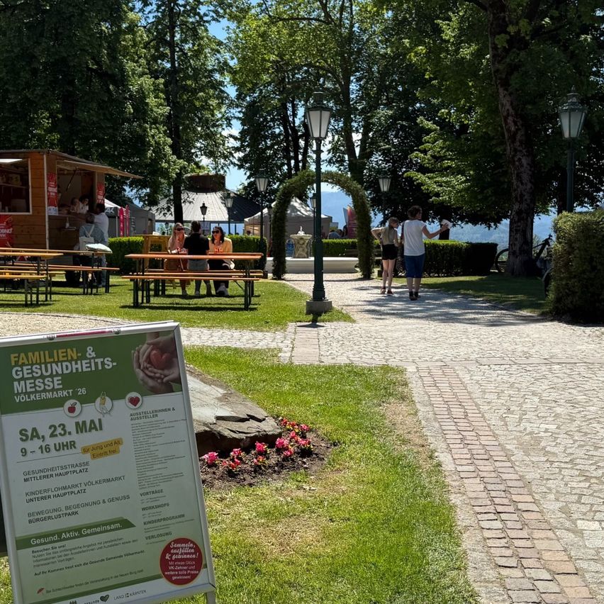 Ein Schild steht in einem Park mit Leuten, die an Picknicktischen sitzen. Es gibt Bäume und Laternen im Hintergrund. Ein Weg führt zu einem Grasbereich.