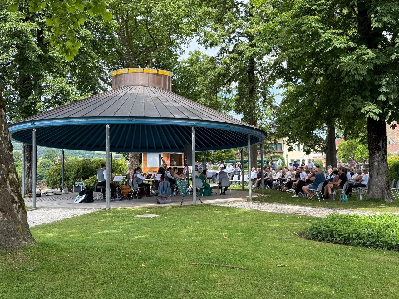 Ein Orchester spielt unter einem Pavillon an einem sonnigen Tag. Viele Menschen sitzen um die Musiker herum und genießen das Open-Air-Konzert. Bäume umgeben das Areal und spenden Schatten.