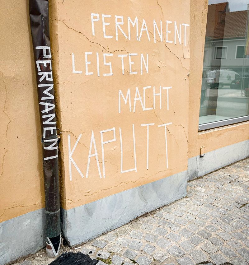 Graffiti an der Gebäudeseite, das 'Permanent leisten macht kaputt' steht. Ein schwarzer Rohr ist an der Wand befestigt.