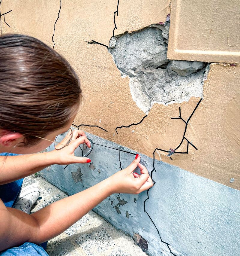 Eine Frau zeichnet mit einem Marker Linien auf eine beschädigte Wand. Sie trägt eine Brille und hat roten Nagellack.
