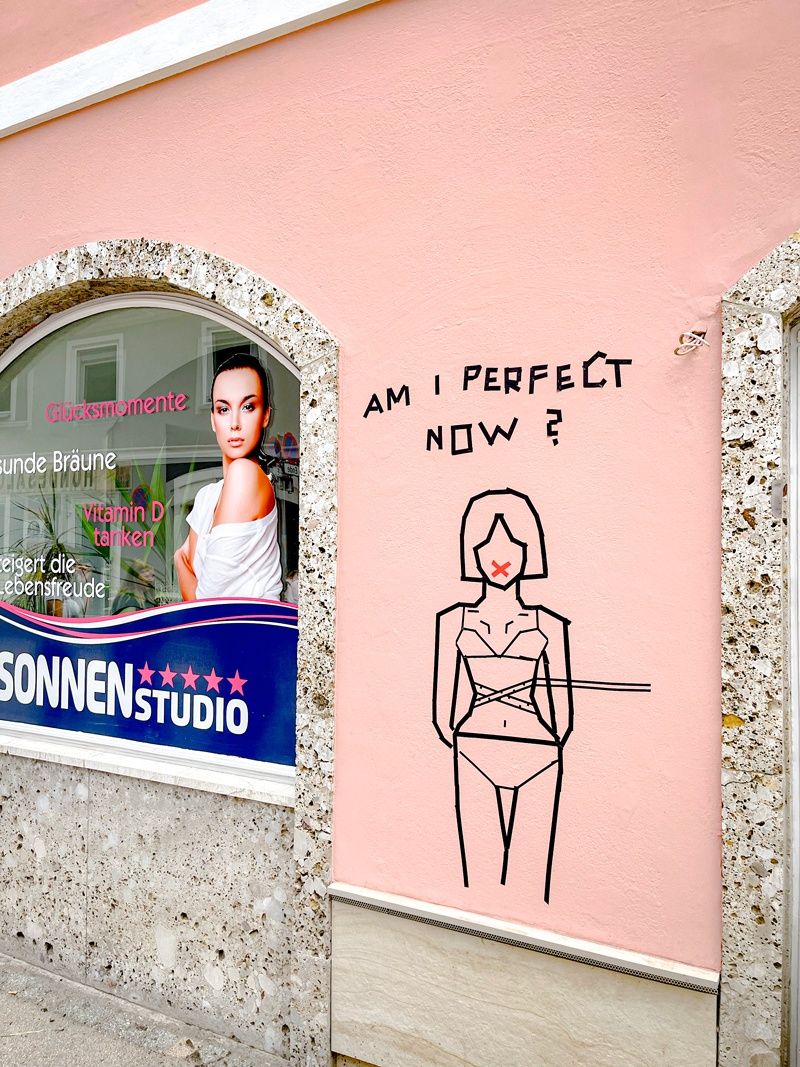 Eine rosafarbene Wand mit einer Zeichnung einer Frau mit einem X auf ihrem Gesicht und den Worten 'Bin ich perfekt jetzt?'. Daneben zeigt ein Fenster ein Plakat einer Frau mit den Worten 'Glücksmomente' und 'Vitamin D tanken'.