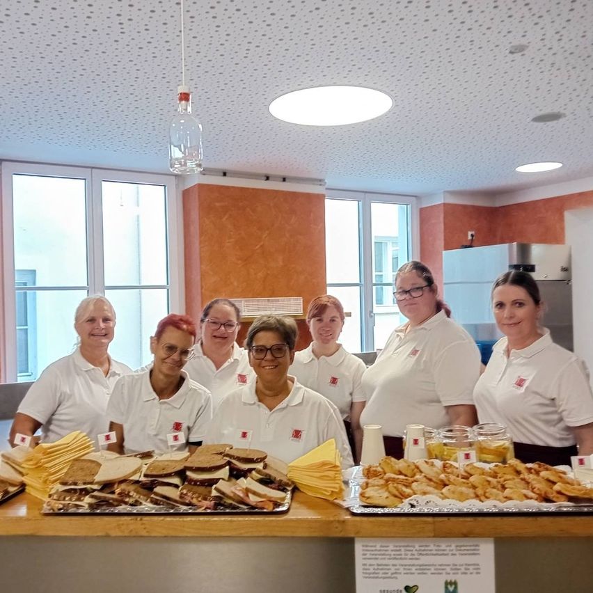 Eine Gruppe von Frauen in weißen Hemden steht hinter einem Tisch, der mit Sandwiches, Keksen und Getränken gefüllt ist. Der Raum hat große Fenster und eine orange Wand.