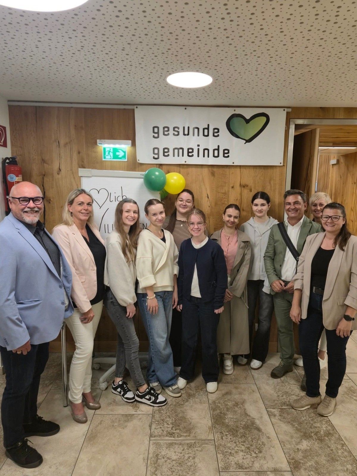 Eine Gruppe von Erwachsenen und jungen Frauen steht zusammen vor einer Holzwand mit einem Banner, das 'gesunde gemeinschaft' steht. Im Hintergrund befinden sich Luftballons und ein Feuerlöscher.