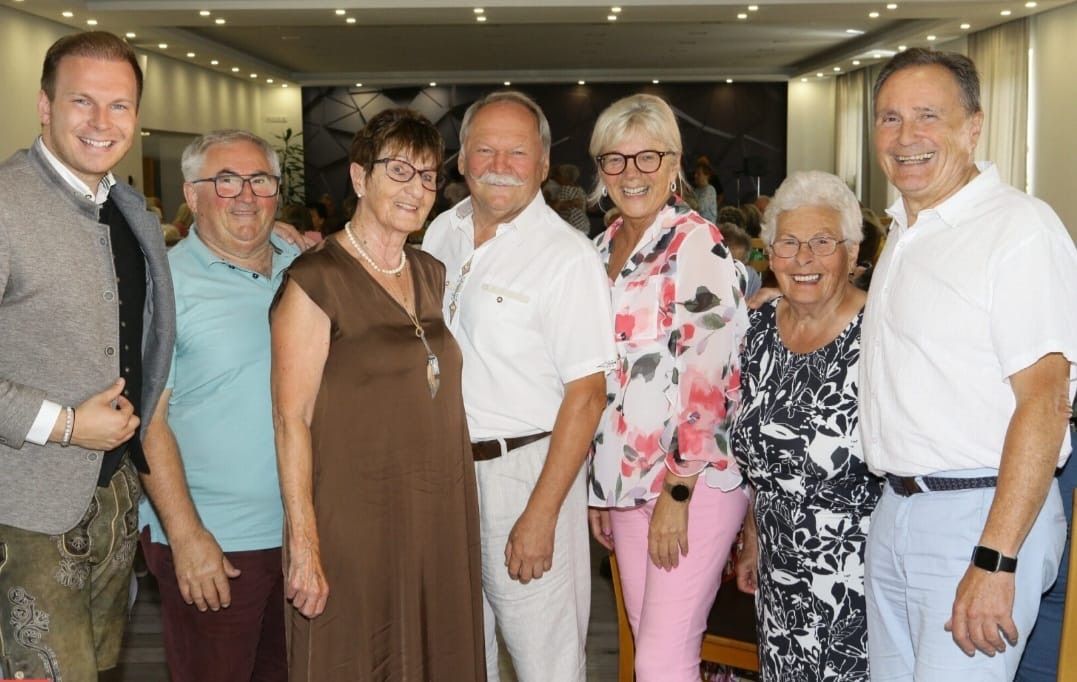 Eine Gruppe älterer Menschen steht eng beieinander, lächelt und posiert für ein Foto. Sie befinden sich in einem gut beleuchteten Innenraum.