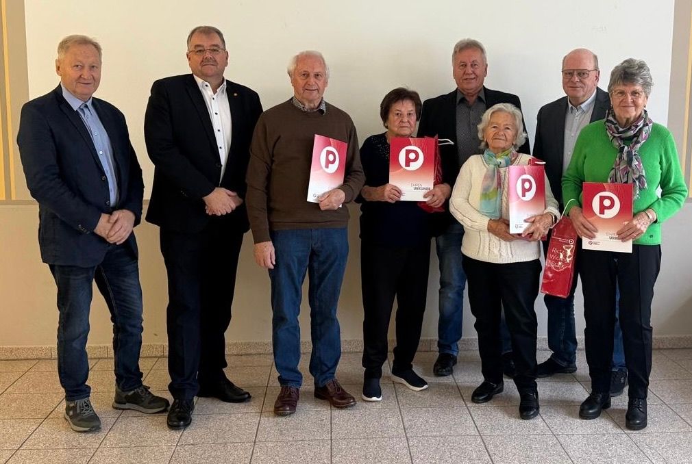 Eine Gruppe von Personen, einige mit Brille, steht in einer Reihe. Sie halten rote Broschüren mit einem 'P' darauf. Der Raum ist weiß und der Boden gefliest.