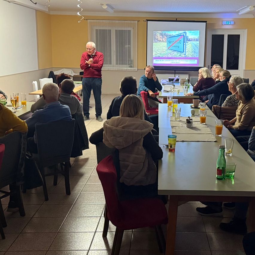 Ein Mann steht vor einer Projektionsleinwand und präsentiert einer Gruppe älterer Erwachsener, die an einem langen Tisch sitzen. Der Raum hat eine gelbe Wand und ein Fenster mit Vorhängen.