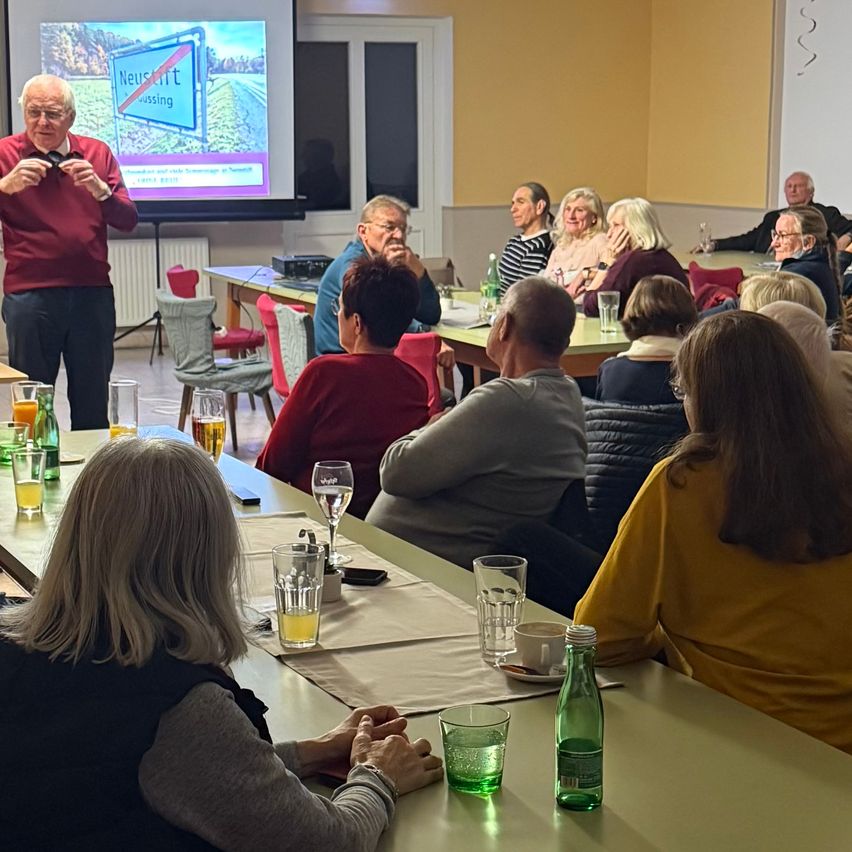 Eine Gruppe von Menschen sitzt an Tischen in einem Raum. Ein Mann mit Brille spricht zu ihnen, und eine Projektionsleinwand zeigt ein Bild hinter ihm.