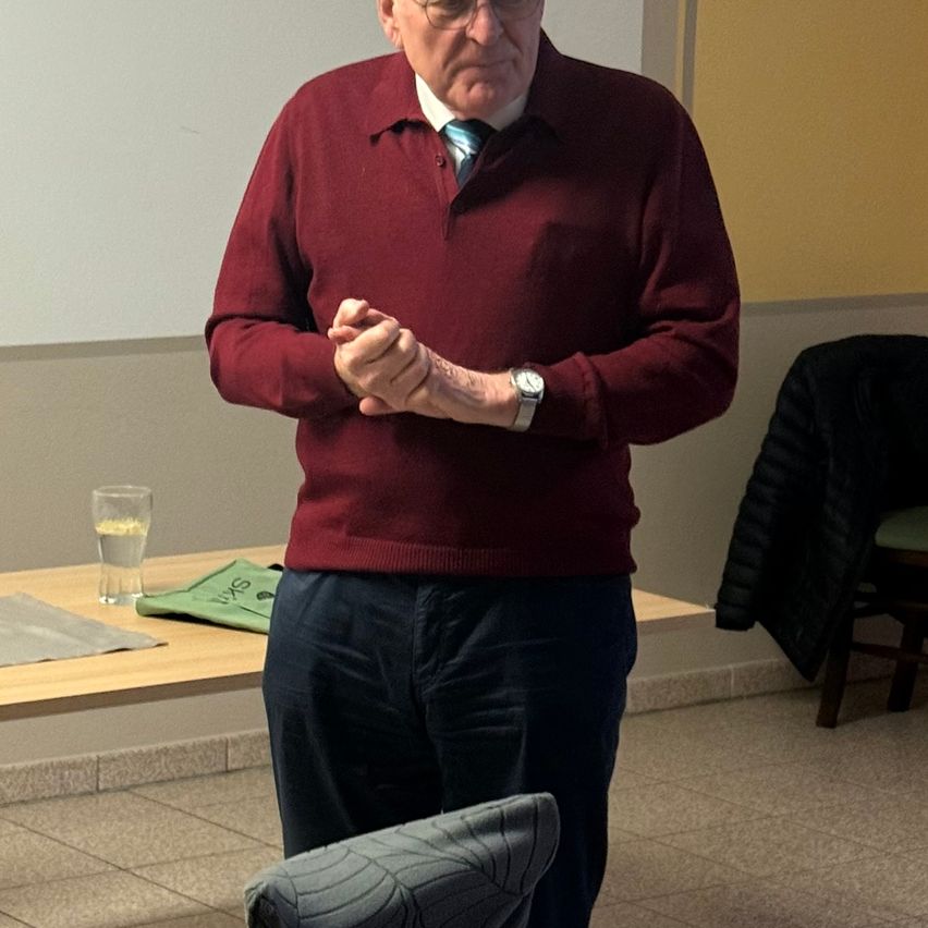 Ein älterer Mann in einem burgunderroten Pullover und Krawatte steht in einem Raum, möglicherweise bei einer Präsentation. Er hält seine Hände vor sich verschränkt. Hinter ihm befindet sich ein Tisch mit einem Glas Wasser und einem grünen Tuch.