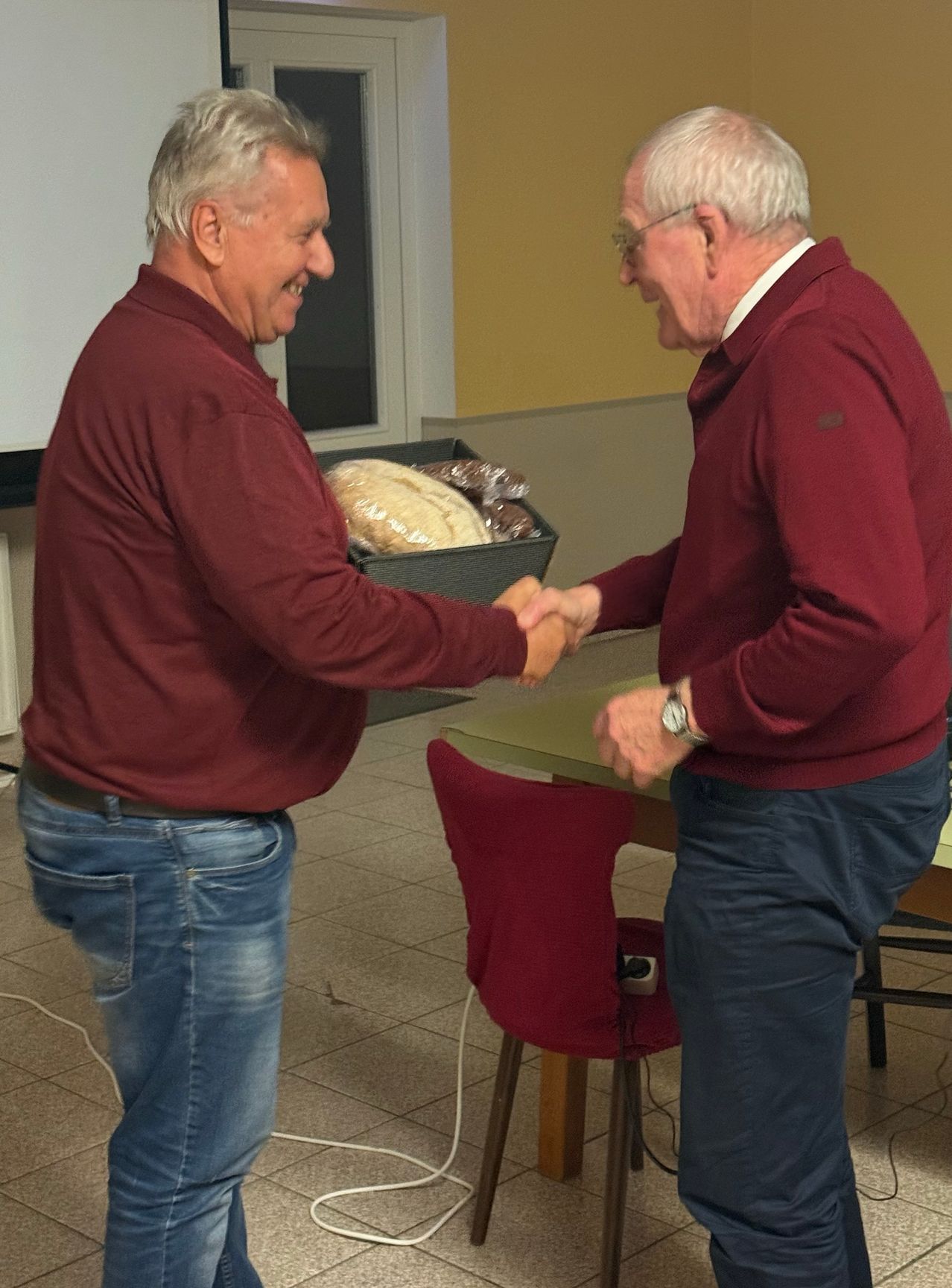 Zwei Männer in weinroten Pullovern schütteln sich die Hände, einer hält eine Brotbox, in einem Raum mit gelber Wand und rotem Stuhl.