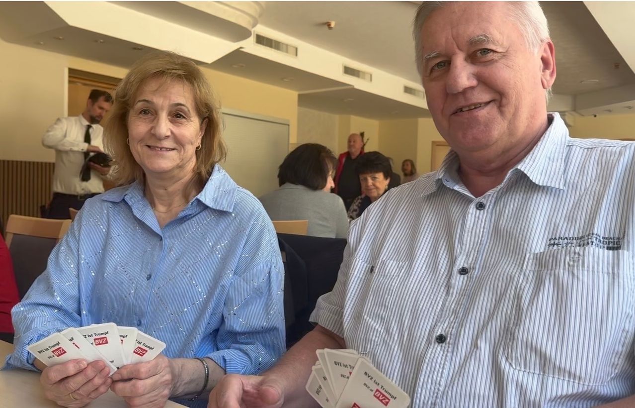 A smiling elderly woman and man hold cards in their hands in a room with other people in the background.