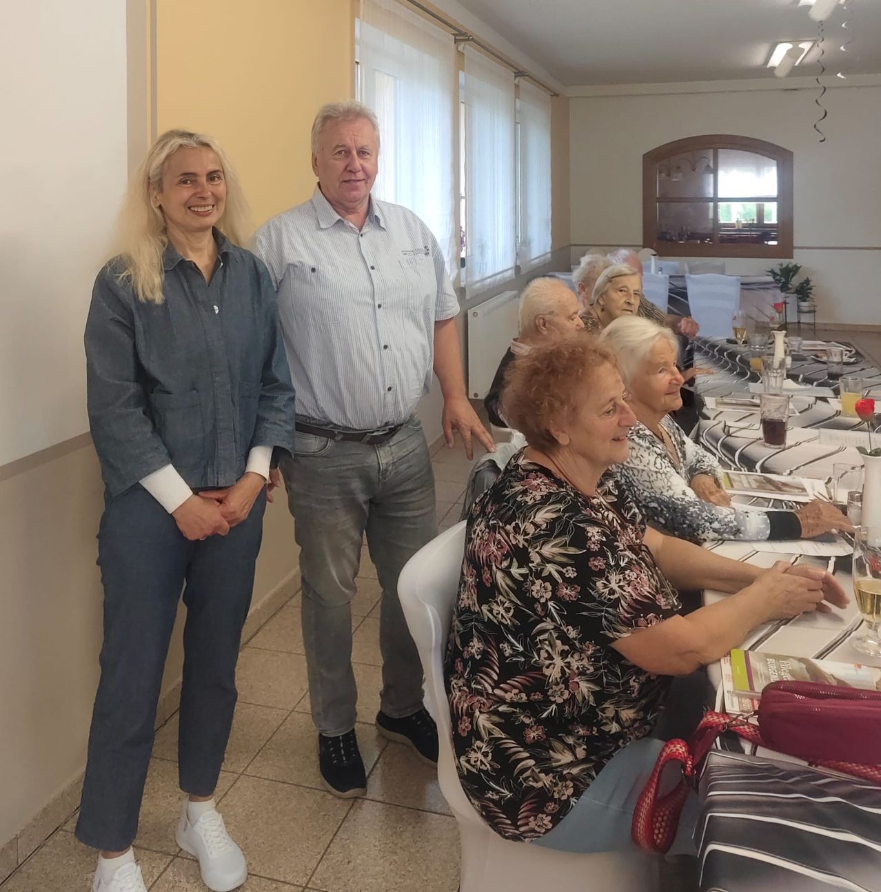 Ein Mann und eine Frau stehen vor einem Tisch mit älteren Frauen, die sitzen. Sie scheinen sich bei einer Zusammenkunft zu befinden, mit Getränken und Snacks auf dem Tisch.
