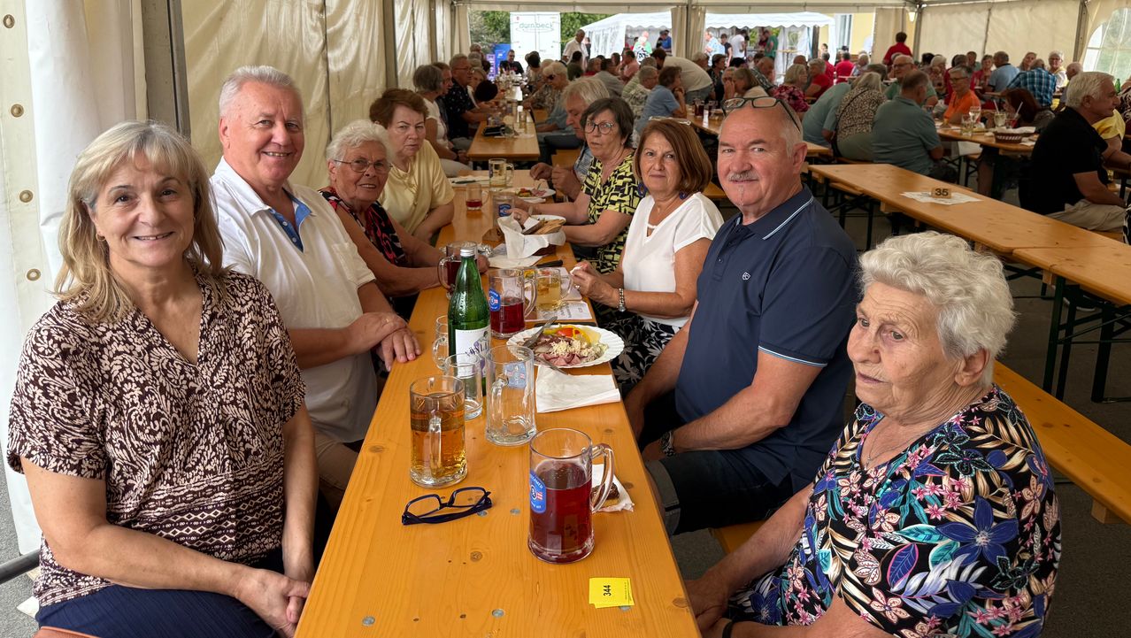 A group of elderly people are sitting at a long table in a tent, smiling and posing for a photo. They are surrounded by plates, glasses, and bottles. The table is filled with food and drinks. In the background, there are other people sitting at tables and tents.