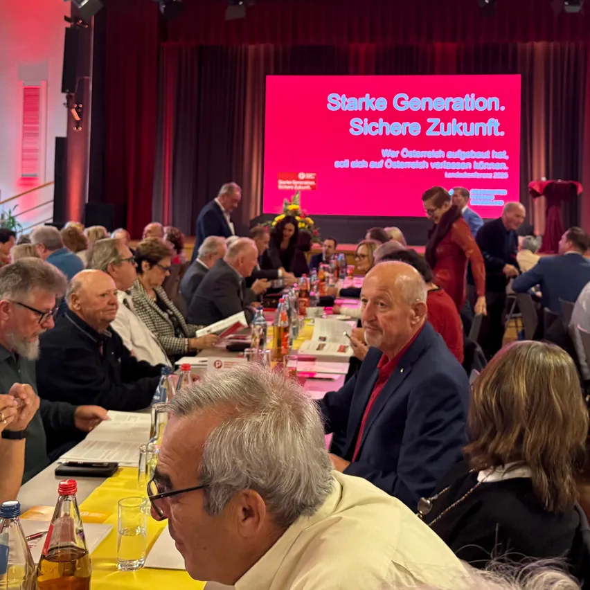 Landeskonferenz 2023: Männer und Frauen sitzen an Tischen, im Hintergrund eine Leinwand mit Text