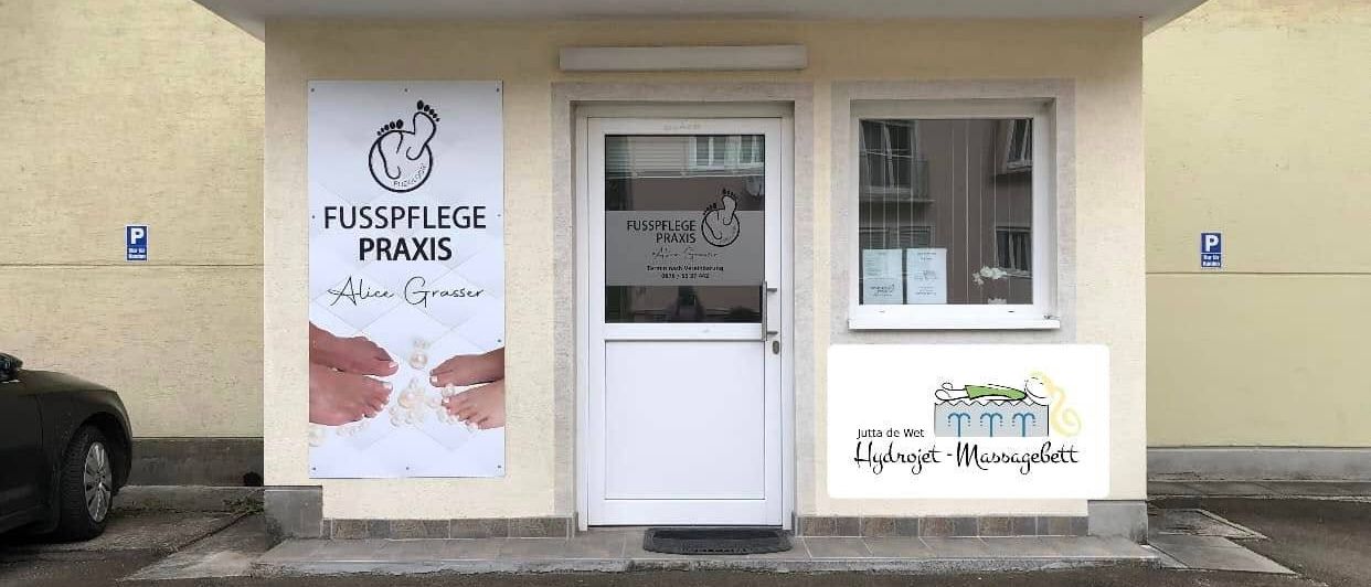 Ein Gebäude mit einer weißen Tür und einem Schild, das 'Fusspflege Praxis' mit einem Logo und dem Namen Alice Grasser besagt. Es gibt auch ein Fenster mit Blick nach draußen.