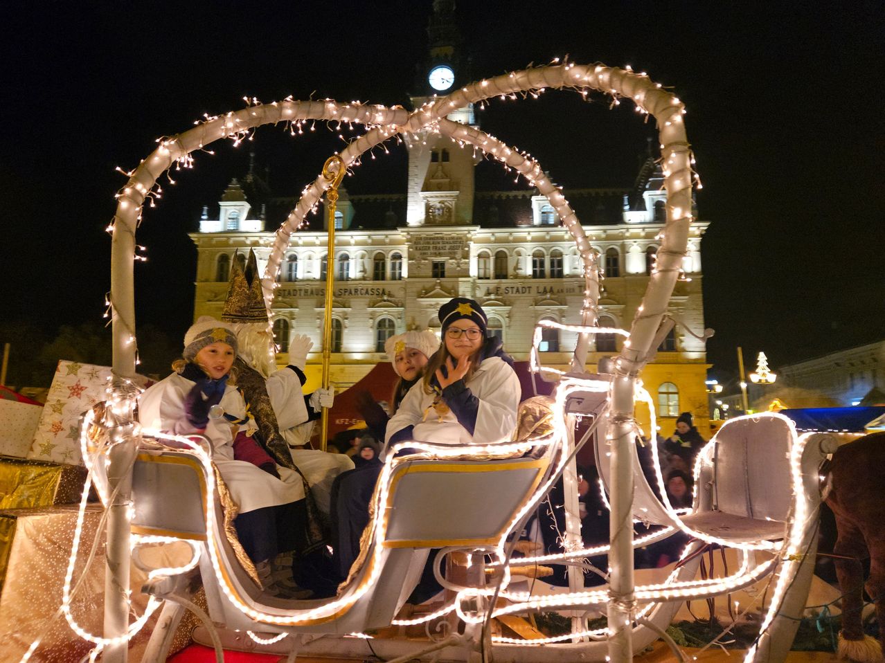 A festive carriage with people dressed in winter costumes sits in front of a building with a clock tower. The carriage is decorated with lights and a golden staff. People are standing behind the carriage.