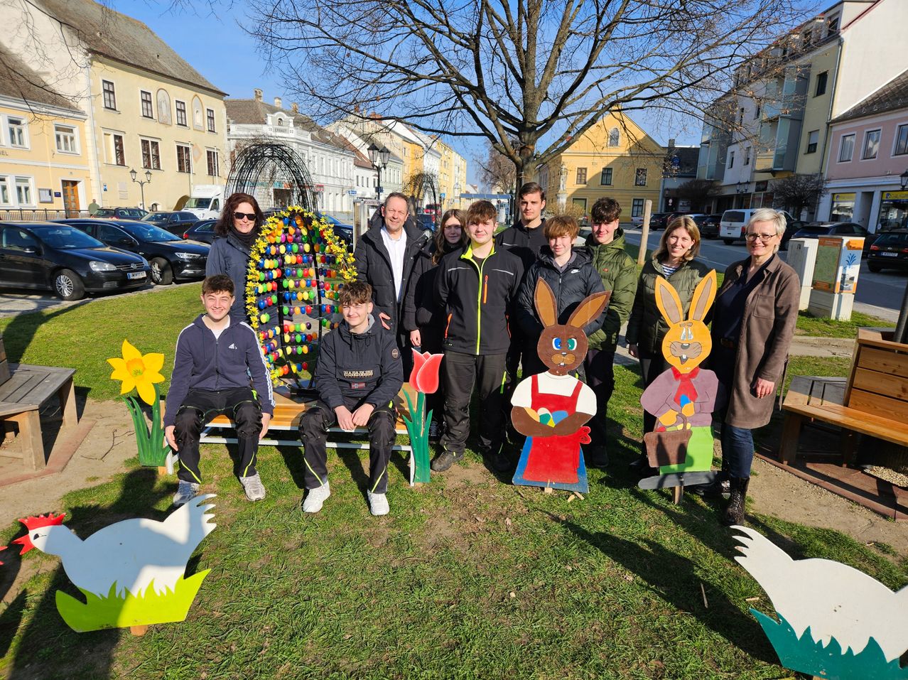 Eine Gruppe von Menschen steht in einem Park in der Nähe von Osterdekoration. Einige sitzen auf einer Bank, andere stehen hinter Hasenausschnitten. Im Hintergrund befinden sich Gebäude und geparkte Autos.