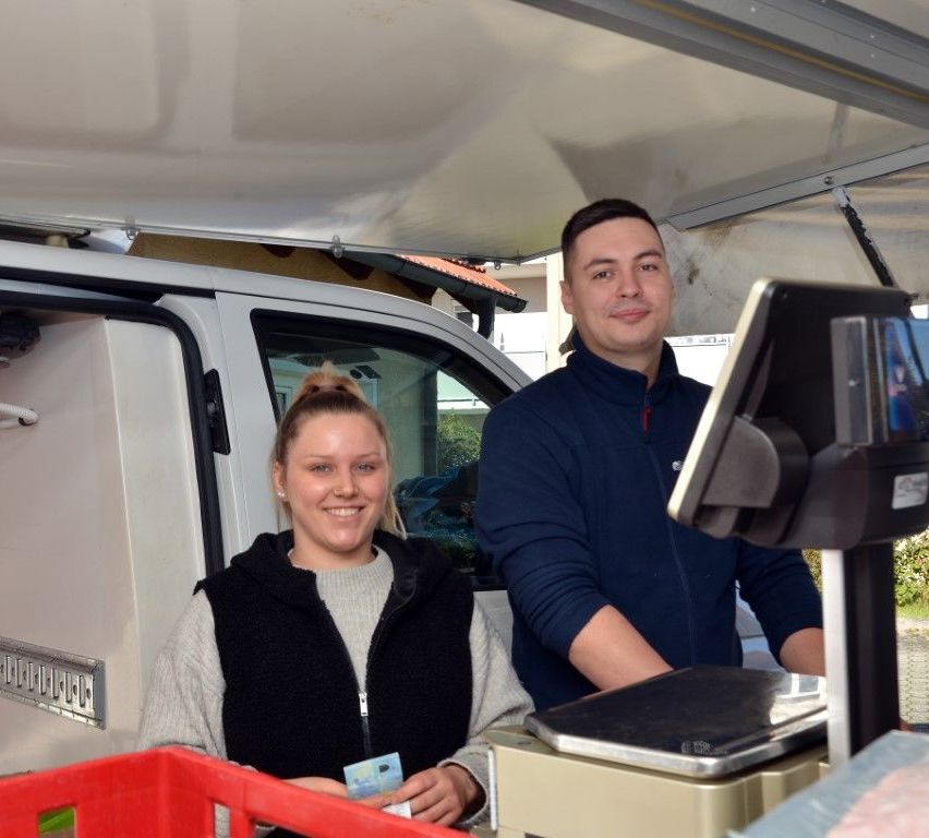 A man and a woman stand in front of a van, smiling. The woman holds a card, and the man has his hand on a scale. The van's door is open, and a red basket is nearby.