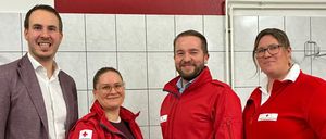 Vier Personen posieren für ein Foto in einem Raum mit roten Kreuzen an der Wand und weißen Fliesen. Sie tragen alle rote Jacken.