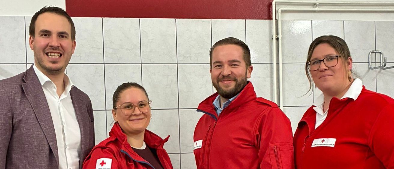 Vier Personen posieren für ein Foto in einem Raum mit roten Kreuzen an der Wand und weißen Fliesen. Sie tragen alle rote Jacken.