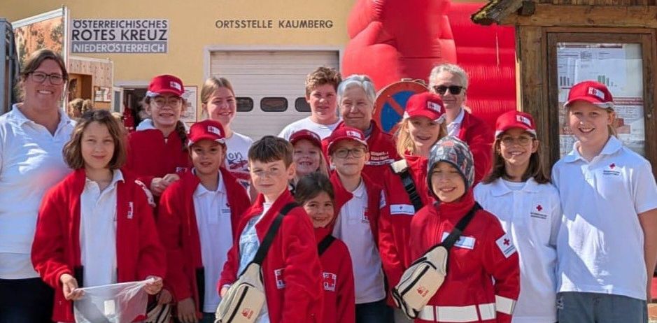 Eine Gruppe von Menschen in roten Jacken und Mützen, einige mit Brillen, stehen vor einem Gebäude. Dahinter befindet sich ein rotes Aufblasobjekt.