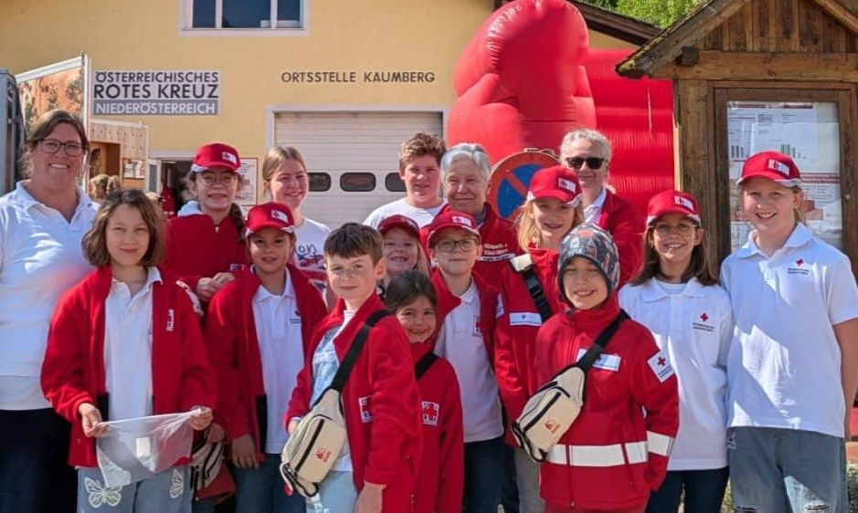 Eine Gruppe von Menschen in roten Jacken und Mützen, einige mit Brillen, stehen vor einem Gebäude. Dahinter befindet sich ein rotes Aufblasobjekt.