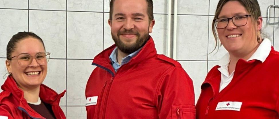 Drei Personen in roten Jacken mit weißen Streifen stehen zusammen. Sie lächeln und scheinen für ein Foto zu posieren.