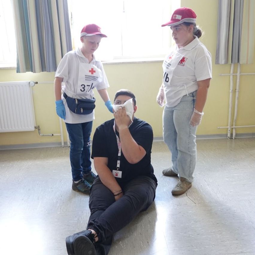 Drei Personen in einem Raum, eine sitzt auf dem Boden und hält ein Tuch vors Gesicht, zwei stehen und tragen weiße Uniformen, eine mit der Nummer 16 und die andere mit 37.