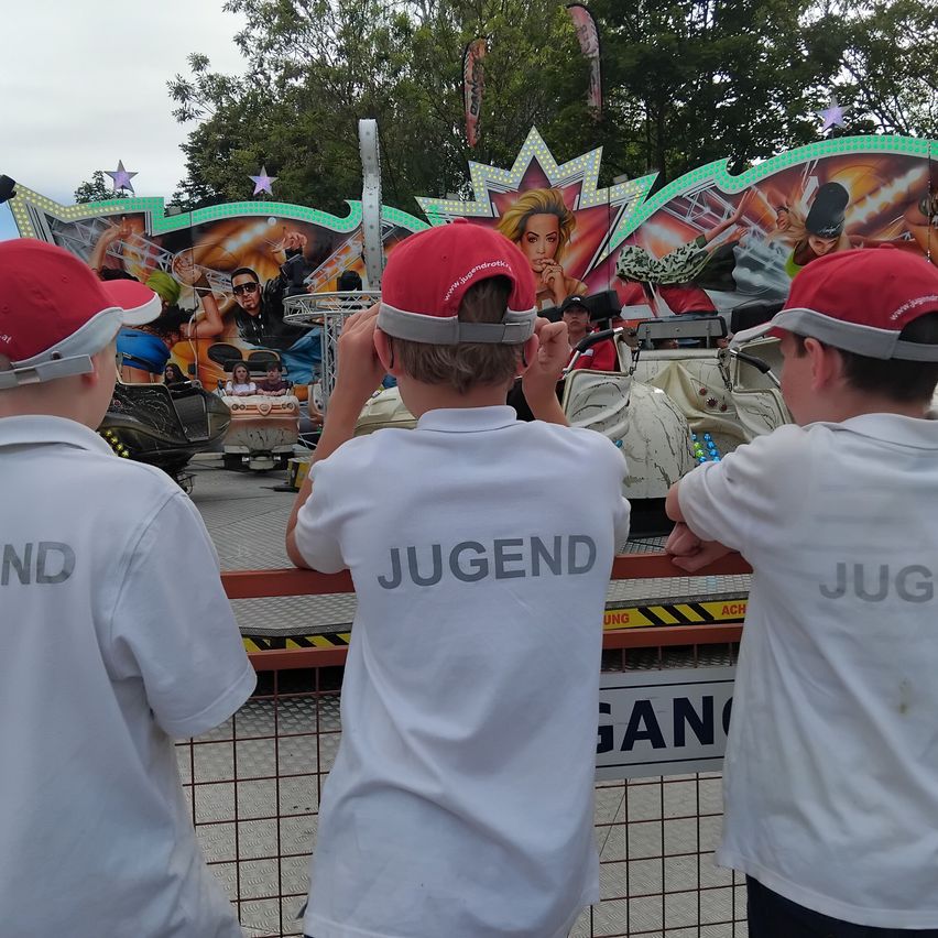 Drei Jungen in weißen Hemden und roten Mützen beobachten eine Drehkarussell in einem Vergnügungspark.