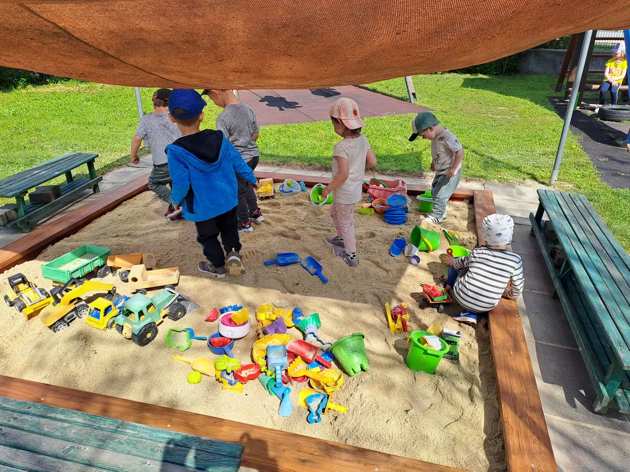 Kinder spielen an einem sonnigen Tag mit Spielzeug in einem Sandkasten, unter einem Baldachin.