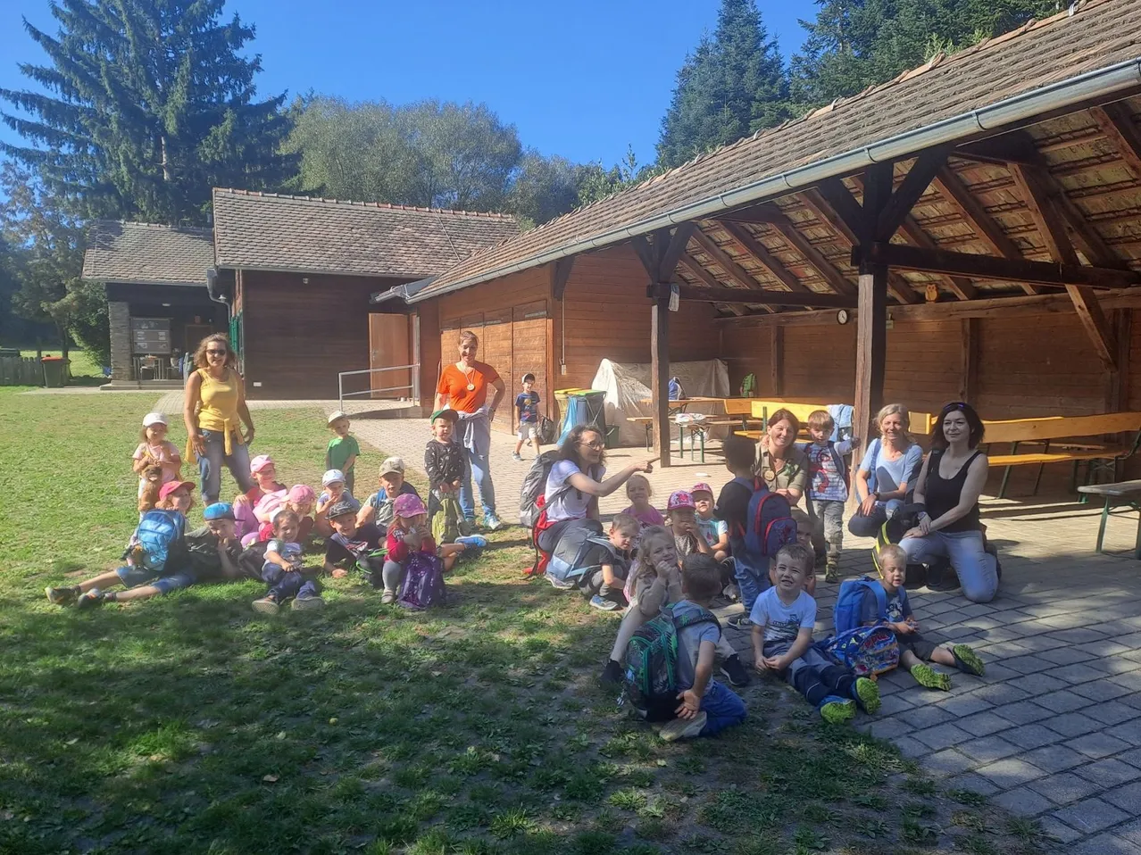 Eine Gruppe von Kindern und Erwachsenen versammelt sich im Freien. Einige sitzen auf dem Rasen, andere stehen. Sie tragen Hüte und Rucksäcke. Hinter ihnen ist ein Holzgebäude mit Dach zu sehen.