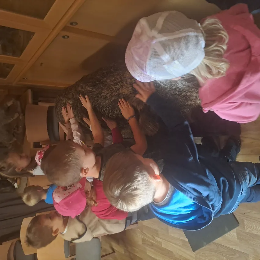 Eine Gruppe von Kindern ist um ein großes Tier, möglicherweise einen Bären, versammelt, mit den Händen darauf. Sie scheinen sich in einem Raum mit Holzböden und Schränken zu befinden.