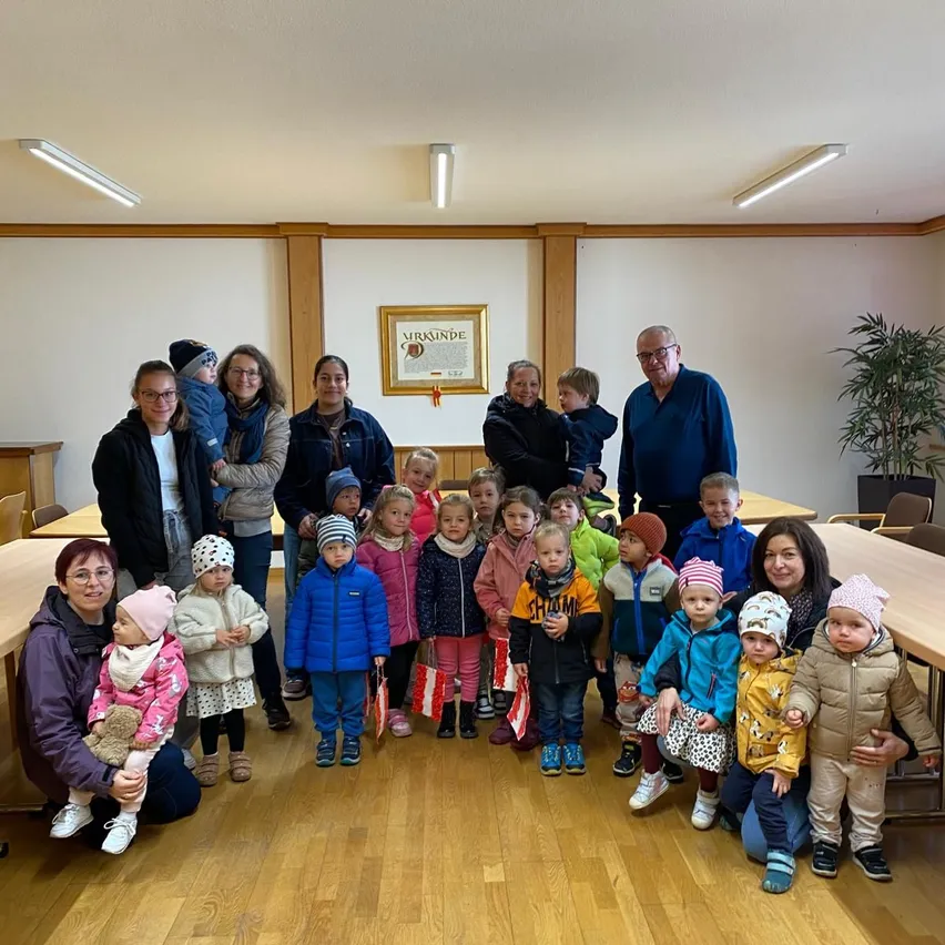 Eine Gruppe von Kindern und Erwachsenen, einige mit Flaggen, posiert für ein Foto in einem Raum mit Holzböden und weißen Wänden. Ein gerahmtes Dokument ist an der Wand angebracht.