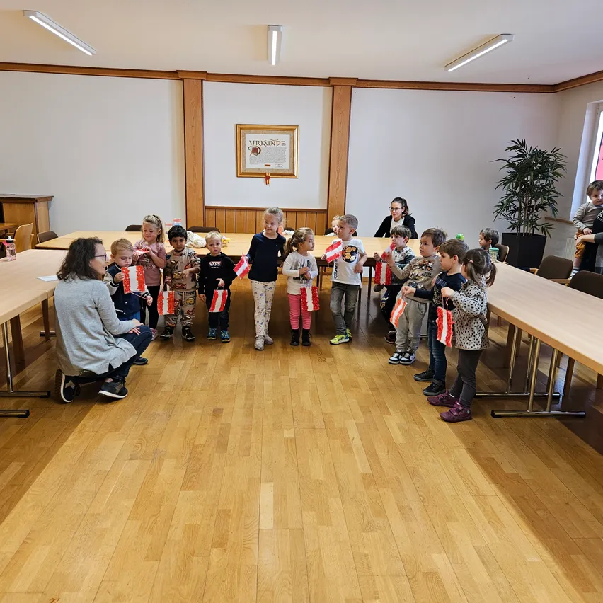 Eine Gruppe von Kindern und Erwachsenen befindet sich in einem Raum mit Holzboden. Sie halten Flaggen und scheinen ein Ereignis zu feiern.