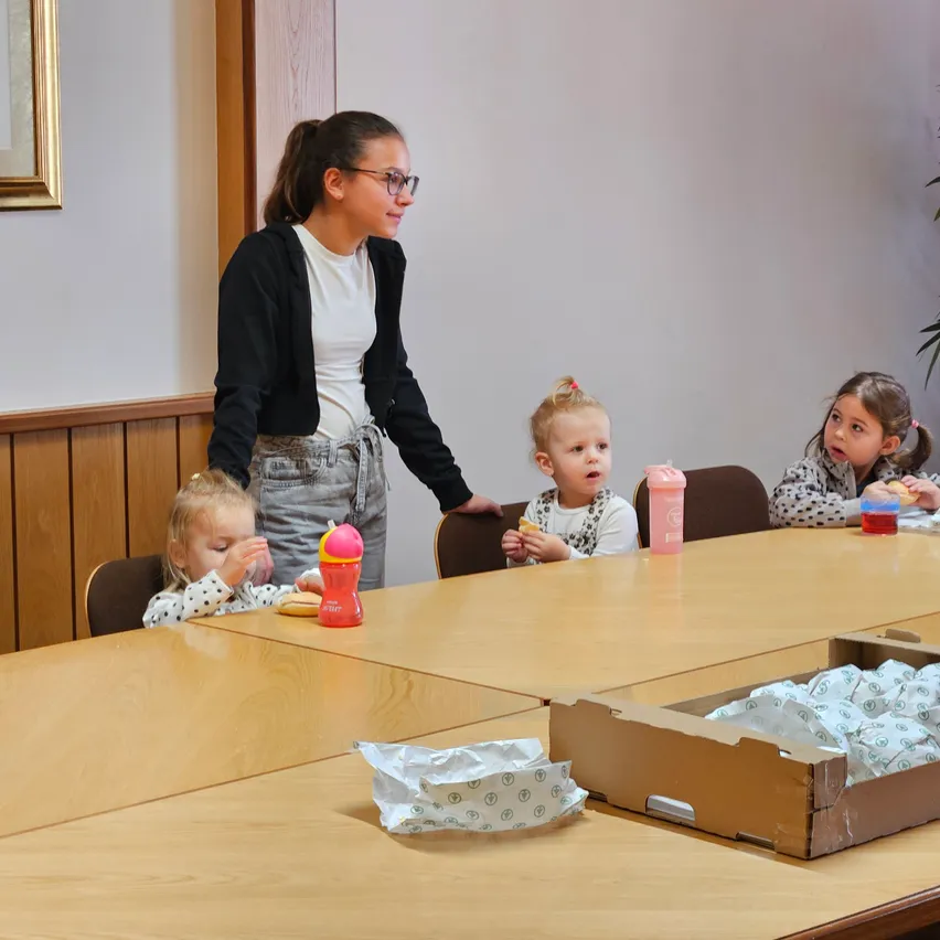 Eine Frau steht neben einem Tisch, an dem vier Kinder sitzen. Die Kinder essen. Die Frau hält eine Flasche. Auf dem Tisch liegen Essen und Getränke. Im Hintergrund befinden sich eine Holzwand und eine Pflanze.