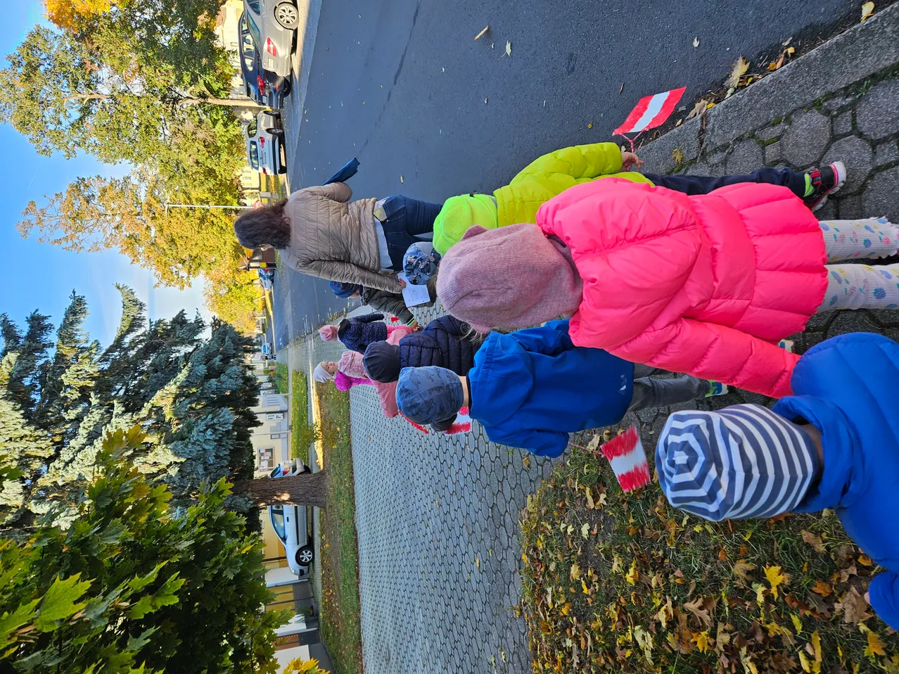 Eine Gruppe von Kindern in Winterjacken und Mützen steht auf einem Bürgersteig, einige halten Flaggen. Eine Frau steht vorne, möglicherweise führt sie sie. Bäume und geparkte Autos sind im Hintergrund.