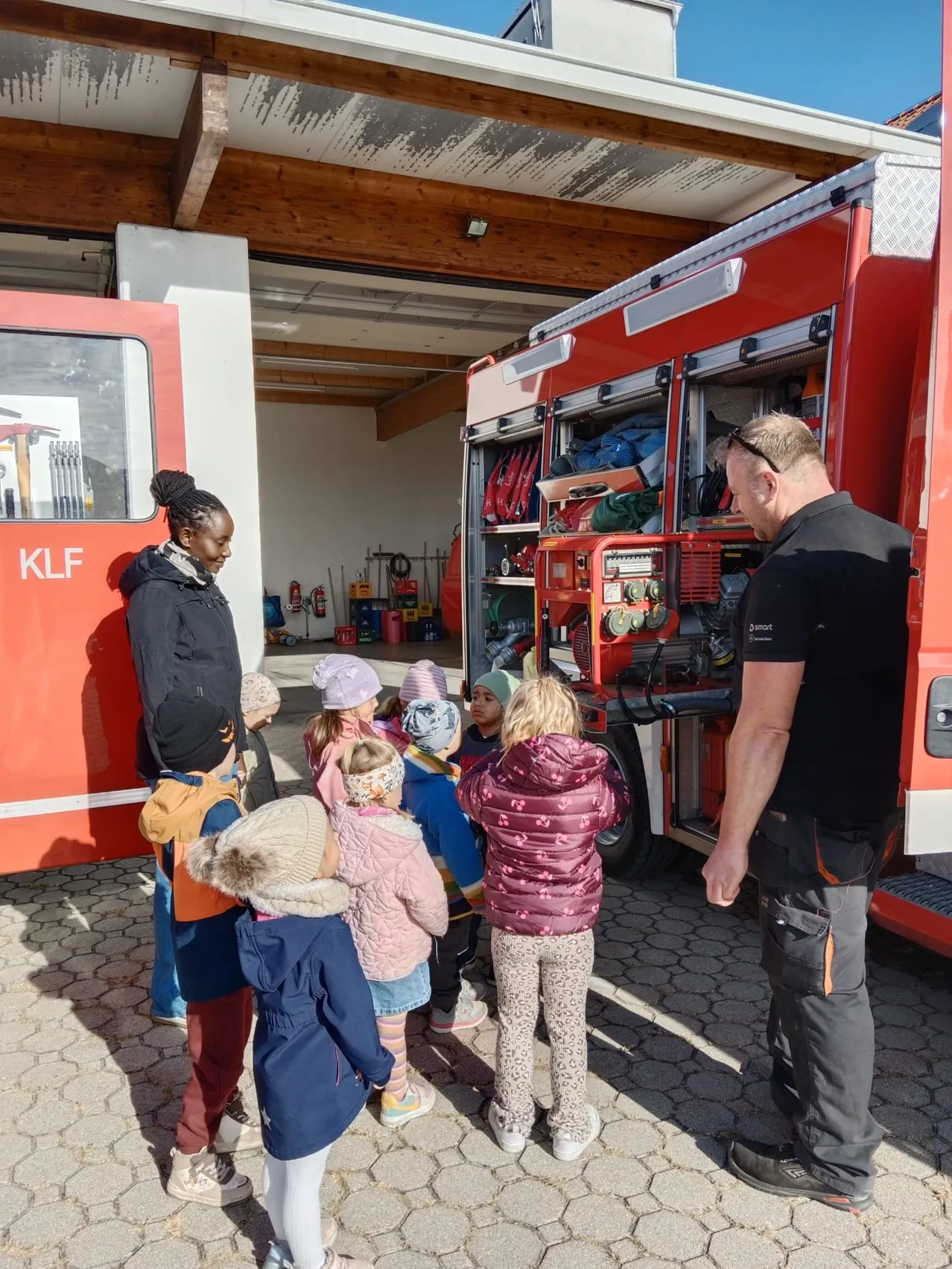 Eine Gruppe von Kindern und zwei Erwachsenen stehen vor einem Feuerwehrauto. Das Feuerwehrauto ist vor einem Gebäude mit einem Holzdach geparkt.