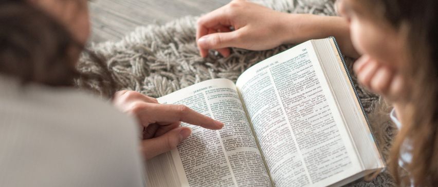 Zwei Personen lesen gemeinsam ein Buch, wobei eine auf den Text zeigt. Das Buch liegt auf einem Teppichboden offen.